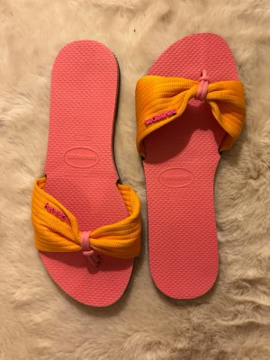 Rosa och orange sandaler Havaianas - Flipflops från Havaianas med rosa sula och breda orange tygband knutna framtill. Märkesnamnet är broderat i rosa på banden. Perfekta för sommaren. 