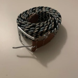 Randigt flätat bälte med läderdetaljer - Snyggt flätat bälte i svart, vitt och grått med randigt mönster. Bältet har silverfärgat spänne och bruna läderdetaljer vid ändarna. Perfekt accessoar för att lyfta din outfit.