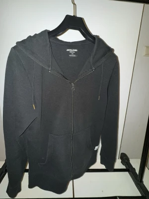 Svart zip hoodie från Jack & Jones - Snygg svart hoodie från Jack & Jones med dragkedja framtill, två stora fickor och justerbar huva med snören. Tillverkad i mjukt bomullsmaterial och har en liten vit logolapp på ena fickan. Perfekt för en avslappnad och stilren look.