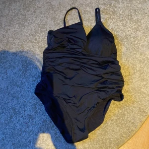 Svart baddräkt från Lindex, M - Stilren svart baddräkt från Lindex i storlek M. Tillverkad av återvunnen polyamidblandning, med tunna axelband och lätt vadderade kupor. Snygg rynkning framtill ger en smickrande passform. Perfekt för stranden eller poolen. Endast använd en gång då jag hade simprov i skolan 