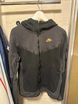 Svart Nike tech fleece - Snygg svart hoodie från Nike med hel dragkedja framtill och guldig Nike logga på bröstet.