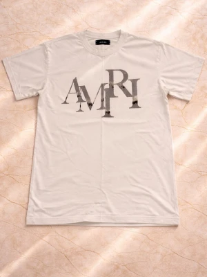 Vit AMIRI t-shirt med tryck - Säljer en vit t-shirt från AMIRI med stort grafiskt tryck i svart och grått på bröstet. Klassisk rund hals och korta ärmar. Tillverkad i mjuk bomull för en skön och avslappnad känsla. Perfekt till jeans eller shorts för en clean look passar även M