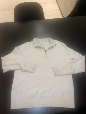 Beige halvzip från These Glory Days - Snygg beige stickad tröja från These Glory Days med halvzip och hög krage. Tröjan har långa ärmar och ribbade muddar vid ärmslut och nederkant. Perfekt för lager på lager och enkel att matcha med jeans eller chinos.