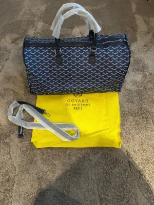 Blå resväska med mönster - Snygg blå resväska med klassiskt vitt mönster och svarta detaljer. Väskan har två handtag och en avtagbar axelrem, samt dragkedja upptill. Materialet är canvas med läderdetaljer. Perfekt för resor och weekendtrips.