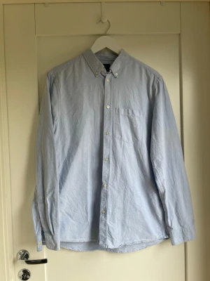 Ljusblå skjorta med knappar - Klassisk ljusblå skjorta med button down-krage och bröstficka. Skjortan har långa ärmar och vita knappar framtill. Perfekt till både jeans och chinos för en clean look.