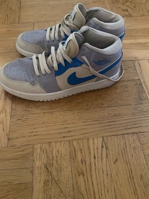 Nike Air Jordan 1 Mid blå/vit - Snygga Nike Air Jordan 1 Mid sneakers i blått, vitt och ljusgrått. Klassisk siluett med vit sula, blå swoosh och detaljer samt perforerad tåbox. Ovandelen är i läder och textil, med vadderad krage för extra komfort. Perfekta för dig som gillar streetwear och ikoniska sneakers.