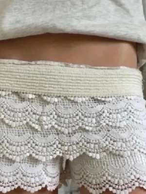 Vita spets-shorts med volang - Säljer ett par vita shorts med bred resår i midjan och flera lager volangig spets. De har en romantisk look med detaljerad spets och scallop-kant. Perfekta för varma dagar och ger en söt och trendig vibe. Köpta utomlands!