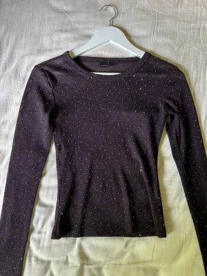 Svart långärmad topp med glitter - Snygg svart långärmad topp med diskreta glittriga prickar över hela tyget. Figurnära passform och rund halsringning. Perfekt att styla med jeans eller kjol för en trendig look.