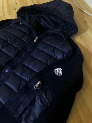 Mörkblå dunjacka från Moncler - Sitter som en Large!Snygg mörkblå cardigan från Moncler med huva och dragkedja framtill. Jackan har quiltade partier, en ficka med dragkedja på ärmen och Moncler-logga på bröstet. Materialet är en mix av dun och nylon, perfekt för kalla vinterdagar.