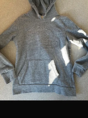 Grå hoodie från Superdry - Snygg grå hoodie från Superdry med broderad logga på bröstet och klassisk känguruficka framtill. Hoodien har huva med dragsko och ribbade muddar vid ärmar och nederkant. Perfekt för en chill och avslappnad stil.