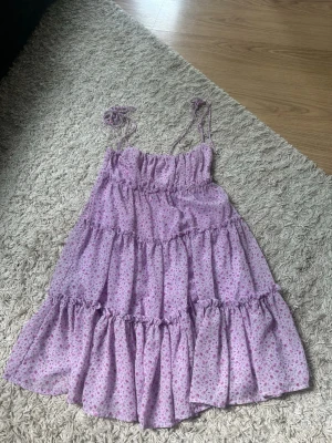 Ljuslila blommig volangklänning - Söt ljuslila klänning med små rosa blommor och volanger. Klänningen har smock upptill och tunna axelband som kan knytas. Perfekt för sommarens alla dagar och ger en romantisk vibe. Materialet känns lätt och luftigt.