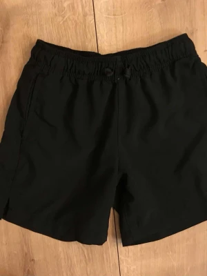 Svarta träningsshorts strl 128/130  - Inga fläckar eller hål, trycket på benet har lite slitage - se bild  Lättviktsshorts i lösare modell med resår i midjan, print på ben och sprund i benslut. Träningsshorts som också passar de yngres aktiva vardag.