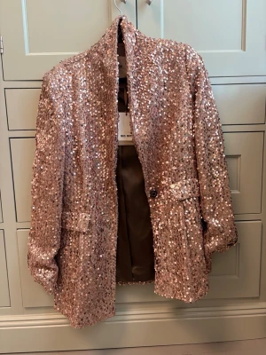 Rosa paljettkavaj från Mos Mosh - Glittrig rosa kavaj från Mos Mosh täckt med paljetter. Kavajen har en rak passform, två fickor framtill och ett knappstängning. Fodrad insida för extra komfort. Perfekt för dig som vill sticka ut och addera lite extra bling till din outfit.
