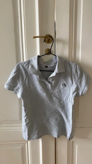 Grå pikétopp med krage och knappar - Klassisk grå pikétopp från H&M med korta ärmar, krage och två knappar framtill. Toppen har en liten broderad logga på bröstet och är tillverkad i mjuk bomull. Perfekt till jeans eller kjol för en avslappnad look.