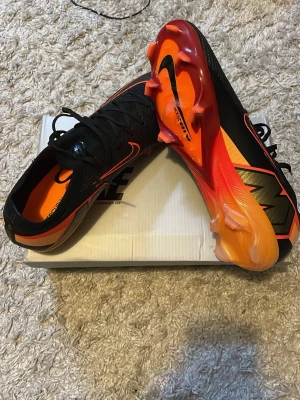 Nike Mercurial fotbollsskor - Säljer ett par Nike Mercurial fotbollsskor storlek 43 i svart och orange med snygga detaljer och en cool sula i orange och gul. Skorna har låg profil, snörning och är tillverkade i syntetmaterial för bästa grepp och känsla på planen. Perfekta för dig som vill sticka ut på fotbollsplanen. Aldrig använda på grund av att dom inte passade mig. Hör av er om ni vill ha fler bilder!