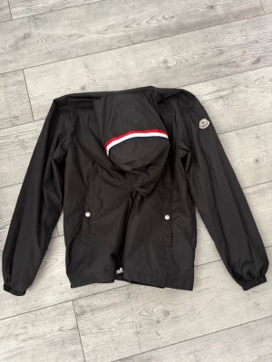 Svart vindjacka från Moncler - Snygg svart vindjacka från Moncler med huva som har röd, vit och mörkblå rand. Jackan har dragkedja framtill, två fickor med tryckknappar och Moncler-logga på ärmen. Perfekt för blåsiga dagar och enkel att matcha till olika outfits. Pris kan alltid diskuteras😁