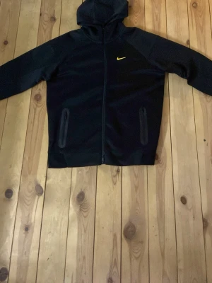 Svart Nike nocta hoodie - Snygg svart hoodie från Nike med dragkedja och huva. Den har två fickor med dragkedja framtill och en gul Nike-logga på bröstet. Perfekt för dig som gillar sportig och stilren design. Materialet känns mjukt och bekvämt. True the size! Priset är fast!