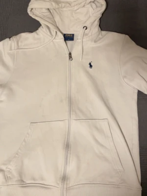 Vit hoodie från Polo Ralph Lauren - Snygg vit hoodie från Polo Ralph Lauren med dragkedja framtill och klassisk blå logga på bröstet. Hoodien har huva med dragsko och stora fickor framtill. Tillverkad i mjukt bomullsmaterial som känns skönt mot huden. Perfekt för en avslappnad och stilren look.