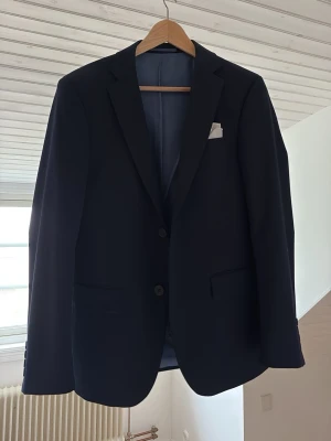 Mörkblå klassisk kavaj - Stilren mörkblå kavaj med två knappar framtill och klassiska slag. Kavajen har två fickor med lock och en bröstficka med vit näsduk. Fodrad insida och enkel, tidlös design som passar till många tillfällen.