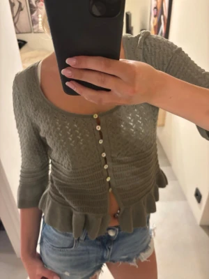 Olivgrön stickad blus med volang - Supersöt olivgrön stickad blus med volangkant och trekvartsärm. Blusen har dekorativa knappar framtill och en croppad passform🌸
