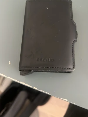 Svart Secrid plånbok i läder - Minimalistisk svart plånbok från Secrid i slätt läder. Plånboken har en kompakt design med präglad logga på framsidan och smart knappfunktion på sidan för enkel åtkomst till kort. Perfekt för dig som gillar stilrena accessoarer.