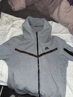 Nike Tech Fleece Hoodie - Säljer en grå hoodie från Nike med hel dragkedja och svart Nike-logga på bröstet. Tröjan har huva, svarta detaljer och en snygg rand över bröstet. Perfekt för chill eller träning. Sitter skönt och har en modern look. Fraktas endast och köparen står för frakt.