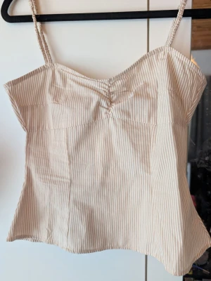 Beige och vit randig linnetopp - Säljer en söt linnetopp med tunna axelband i beige och vitt randigt mönster. Toppen har en lätt rynkning vid bysten och är tillverkad i ett mjukt bomullsliknande material. Perfekt för varma dagar och enkel att styla med jeans eller kjol.