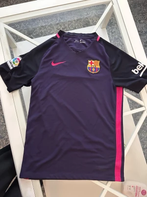 Barça matchtröja lila Nike - Snygg lila FC Barcelona matchtröja från Nike med rosa detaljer och klubbmärke på bröstet. Tröjan har svarta ärmar, La Liga-märke på båda ärmarna och UNICEF-tryck på ryggen. Perfekt för dig som älskar fotboll och vill representera Barça.