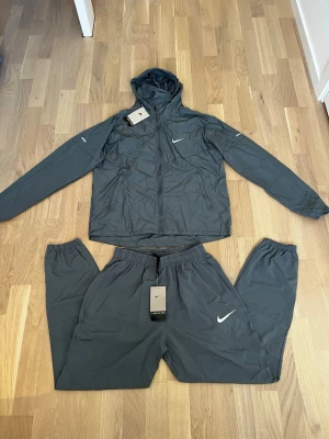 Grå nike tränningset  - Snyggt mörkgrått träningsset från Nike med jacka och byxor. Jackan har huva, dragkedja och två sidofickor samt Nike-logga på bröstet. Byxorna har resår i midjan och Nike-logga på benet. Perfekt för träning eller chill. Materialet är lätt och snabbtorkande.