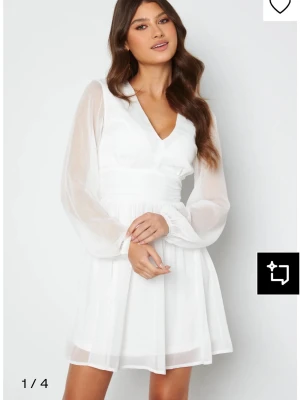 Vit kortklänning med v-ringning - Snygg vit kortklänning med v-ringad hals och transparenta långärmar. Klänningen har markerad midja och lätt utställd kjol. Tillverkad i ett tunt, flowy material som ger en luftig känsla. Perfekt för sommarens alla tillfällen.