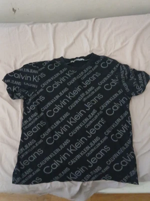 Svart Calvin Klein Jeans t-shirt - Snygg svart t-shirt från Calvin Klein Jeans med stort logomönster över hela plagget. T-shirten har rund halsringning och korta ärmar. Tillverkad i mjuk bomull som känns skön mot huden. Perfekt för dig som gillar streetwear och tydliga loggor.