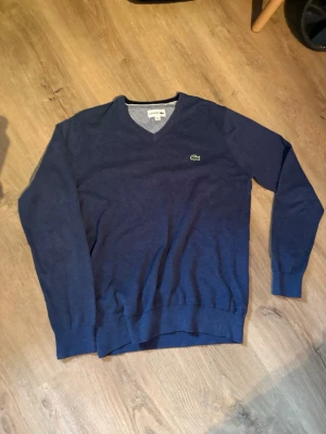 Mörkblå stickad tröja från Lacoste - Snygg mörkblå stickad tröja från Lacoste med klassisk v-ringning och den ikoniska gröna krokodilloggan på bröstet. Tröjan har ribbade muddar vid ärmslut och nederkant. Perfekt för dig som gillar stilrena och tidlösa plagg.