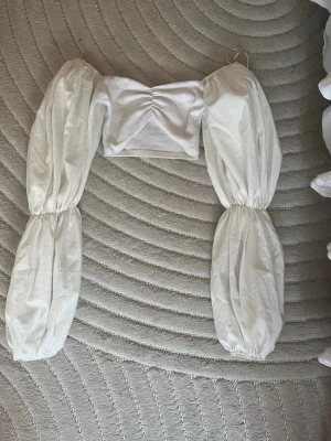 Vit croppad topp med puffärmar - Trendig vit croppad topp med långa puffärmar. Toppen har en offshoulder-design och är tillverkad i ett lätt bomullsliknande material.