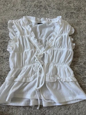Vit volangblus från Gina Tricot - Supersöt vit blus från Gina Tricot i storlek XS. Blusen har volangdetaljer längs ärmhål och halsringning, knytband framtill och smockad midja för en feminin siluett. Tillverkad i ett lätt, strukturerat tyg som känns luftigt och somrigt.