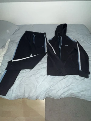 Marinblå BOSS loungewear set med huva - Snyggt svart loungewear set från BOSS med matchande hoodie och byxor. Setet har ljusblå och vita ränder längs ärmar och ben, samt dragsko i midjan på byxorna. Perfekt för chill dagar hemma. Materialet känns mjukt och bekvämt.