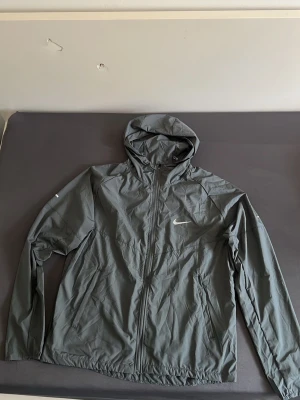 Grå Nike miller - Snygg grå vindjacka från Nike med hel dragkedja och justerbar huva. Jackan har två sidofickor och ett diskret Nike Swoosh på bröstet. Tillverkad i lätt och vindtätt material, perfekt för blåsiga dagar.