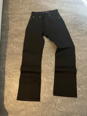 Svarta Bootcut jeans  - Säljer ett par svarta jeans med rak passform och klassisk femficksdesign. Jeansen har bälteshällor och knappgylf. Materialet ser ut att vara kraftig denim, perfekt för en clean och stilren look.