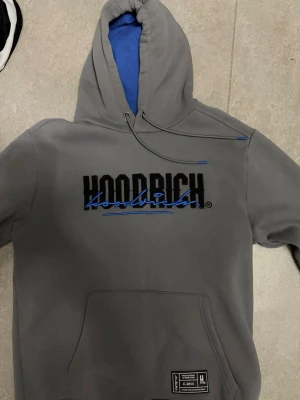 Grå HOODRICH hoodie med blå detaljer - Snygg grå hoodie från HOODRICH med svart och blå logga på bröstet och ryggen. Hoodien har blått foder i huvan, dragsko och stor magficka framtill. Mjuk bomullsblandning och streetwear-vibe, perfekt för dig som gillar stilrena men ändå unika plagg.