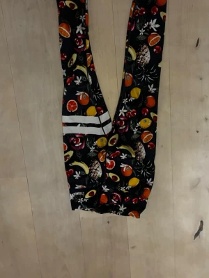 Svarta leggings med fruktmönster - Färgglada svarta leggings från Strong Girls med tropiskt fruktmönster som ananas, banan, apelsin och körsbär. De har vita ränder på ena benet och elastisk midja. Perfekta för dig som gillar unika och lekfulla träningskläder. Skulle säga att de passar xxs-xs 