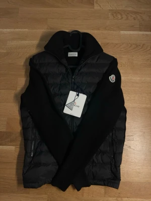  Moncler Cardigan - Snygg svart dunväst från Moncler med stickade svarta ärmar och hög krage. Västen har dragkedja framtill, två sidofickor med dragkedja och Moncler-logga på ena ärmen. Perfekt för lager-på-lager och streetstyle.