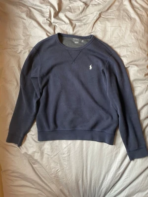 Mörkblå sweatshirt från Polo Ralph Lauren - Mörkblå sweatshirt från Polo Ralph Lauren med klassisk rund halsringning och den ikoniska vita logobroderingen på bröstet. Tröjan har ribbade muddar vid ärmslut och nederkant. Tillverkad i mjuk bomullsblandning för skön komfort. MODELLEN ÄR 181 CM