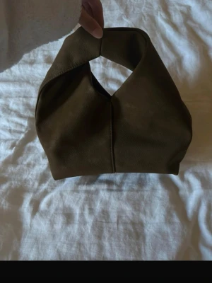 Mörkgrön handväska i mjukt material - Trendig mörkgrön handväska med unik, knutliknande form och kort handtag. Väskan är tillverkad i ett mjukt, slätt material som ger en stilren och modern look. Perfekt accessoar för dig som gillar minimalistisk design och vill sticka ut.