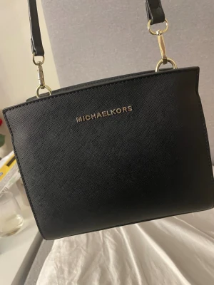 Svart Michael Kors väska - Snygg svart Michael Kors väska med guldfärgade detaljer och justerbar axelrem. Väskan har en fyrkantig form och dragkedja upptill. Perfekt för dig som vill ha en klassisk och trendig accessoar.💞 den är mellanstor