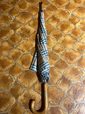 Burberry paraply  - Stilrent paraply med klassiskt beige, svart och rött rutmönster. Paraplyet har ett elegant böjt handtag i trä och metallstomme. Perfekt accessoar för regniga dagar och ger en tidlös look till din outfit.