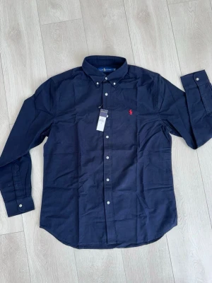 Marinblå skjorta från Ralph Lauren - Snygg marinblå skjorta från Ralph Lauren med klassisk button-down krage och vita knappar. Skjortan har lång ärm och ikonisk röd logobrodyr på bröstet. Perfekt för dig som vill ha en stilren look med premiumkänsla.
