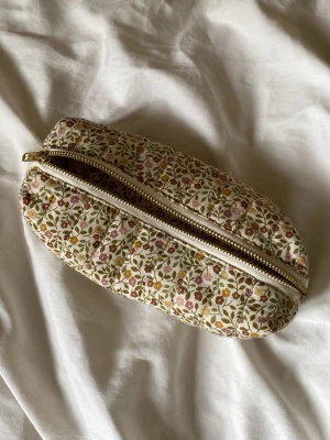 Blommigt quiltat necessärfodral - Söt quiltad necessär med blommigt mönster i grönt, rosa och gult på beige botten. Oval form och dragkedja i guld. Perfekt för småprylar eller smink. Tygmaterial med mjuk känsla och diskret retrostil.