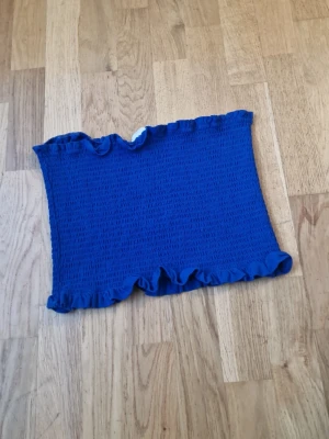 Blå bandeau tubtopp med volangkant - Säljer en blå bandeau tubtopp med smockad struktur och volangkant upptill och nedtill. Toppen är ärmlös och har en stretchig passform som sitter snyggt på kroppen. Perfekt för varma dagar eller som layering-piece.