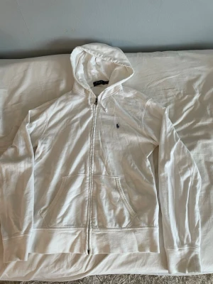 Ralph Lauren Zip up - Vit hoodie från Polo Ralph Lauren med dragkedja och klassisk huva. Den har två stora fickor framtill och en liten broderad logga på bröstet. Materialet är mjukt och bekvämt, perfekt för chill dagar. Enkel och stilren design som funkar till det mesta.