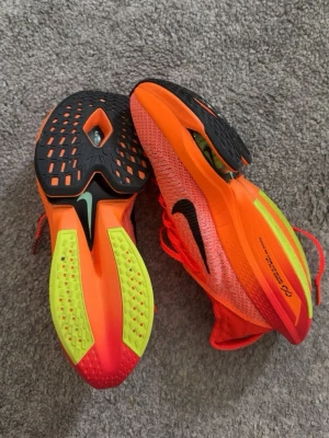 Nike ZoomX Vaporfly Next% 2 löparskor - Säljer ett par Nike ZoomX Vaporfly Next% 2 löparskor i neonorange med svarta detaljer och limegula partier på sulan. Skorna har ventilerande mesh-överdel, responsiv ZoomX-sula och kolfiberplatta för maxad löpkänsla. Perfekta för snabba pass och tävling.