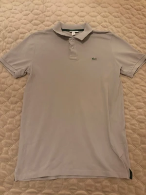 Ljusgrå pikétröja från Lacoste - Klassisk ljusgrå pikétröja från Lacoste med den ikoniska gröna krokodilloggan på bröstet. Tröjan har korta ärmar, krage och två knappar framtill. Perfekt för en clean och stilren look.